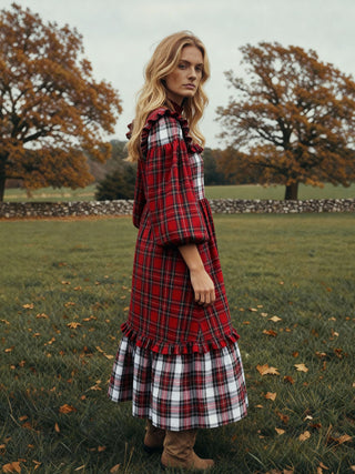 Woman wearing a red plaid dress and coat standing in a field with trees in the background.