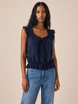 A woman modeling a sleeveless navy blue top with ruffled shoulders, a drawstring neckline, and a button-front bodice, paired with blue jeans against a plain beige studio background.