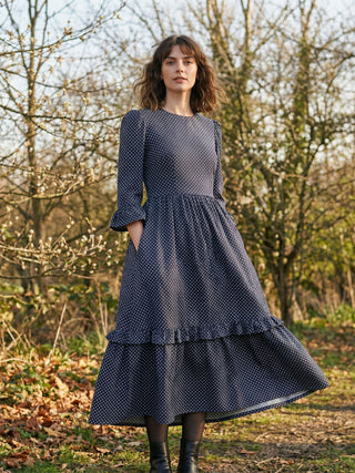 Woman wearing a dark blue polka dot dress standing in a natural setting with trees and grass.