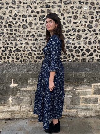A woman wearing a navy blue midi dress with a white floral pattern, standing in front of a rustic stone wall.