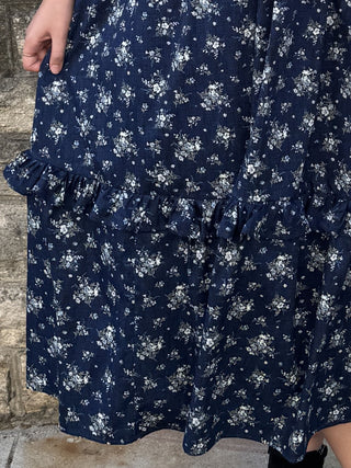 A close-up of a navy blue midi dress with a white floral print and tiered ruffle details, worn by a model standing against a rustic stone wall.