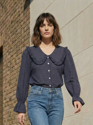 Woman wearing a navy blue polka dot blouse with ruffled collar and high-waisted blue jeans against a brick wall.