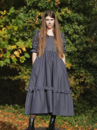 Woman wearing a dark blue polka dot dress standing outdoors with greenery in the background