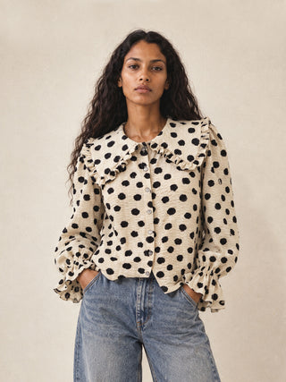A woman wearing a cream-colored long-sleeve blouse with black polka dots, ruffled collar and cuffs, paired with light-wash blue jeans, standing against a neutral beige background.