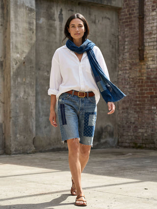 Woman wearing a white shirt, blue denim shorts, and a blue scarf against a brick wall.