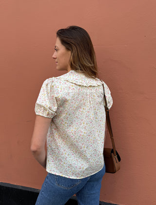A woman shown from the back wearing a cream-colored short-sleeve blouse with a delicate multicolor floral print, paired with blue jeans and carrying a brown shoulder bag against a solid terracotta-colored wall.