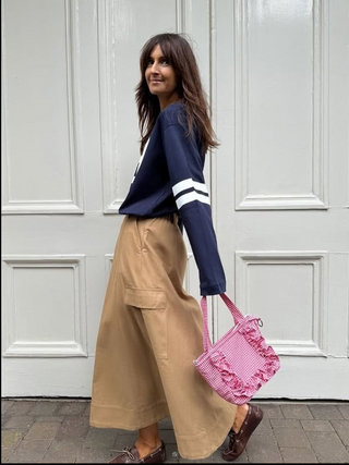 Woman wearing a navy sweater with white stripes and beige pants, holding a pink bag against a white wall.