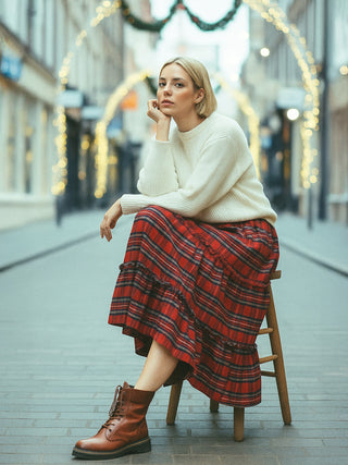 Woman sitting on a stool wearing a white sweater and red plaid skirt with blurred lights in the background