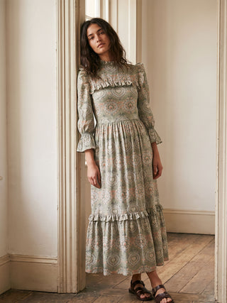 Woman wearing a long, patterned dress standing in a room with wooden flooring and white walls.