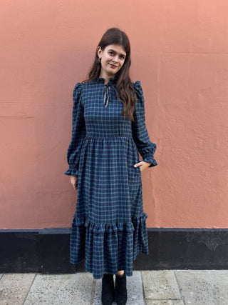 A woman wearing a dark blue and black plaid midi dress with long puff sleeves, a tie-neck detail, and a ruffled hem, standing against a textured terracotta wall.
