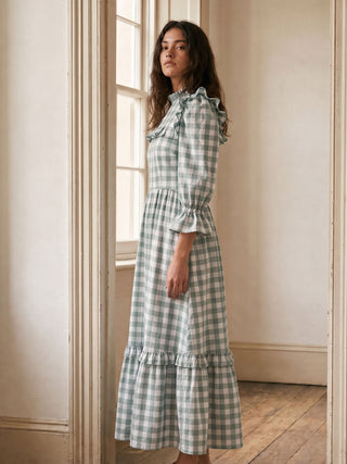 Woman wearing a green and white checkered dress standing in a room with wooden flooring and a window.