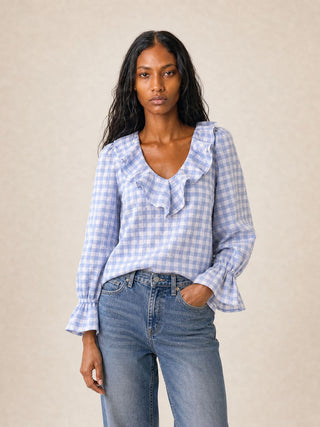 A woman modeling a light blue and white gingham long-sleeve blouse with a ruffled v-neckline and flared cuffs, paired with blue jeans, against a neutral beige background.