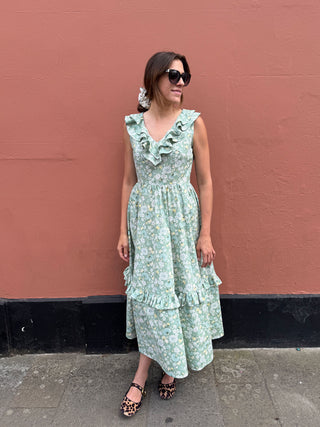 A woman wearing a light green ruffled midi dress with a delicate floral print, standing against a textured terracotta wall.