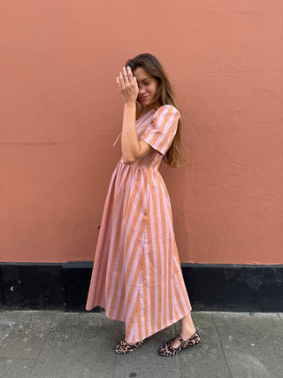 A woman wearing a pink and white vertically striped midi dress with short sleeves, standing against a textured terracotta wall.