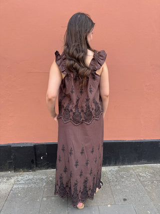 A woman viewed from behind wearing a sleeveless brown skirt and top with black floral embroidery and ruffled shoulder details, standing against a terracotta-colored wall.