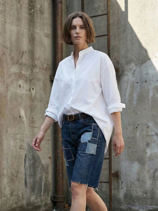 Woman wearing a white shirt and patchwork denim shorts against a concrete wall.
