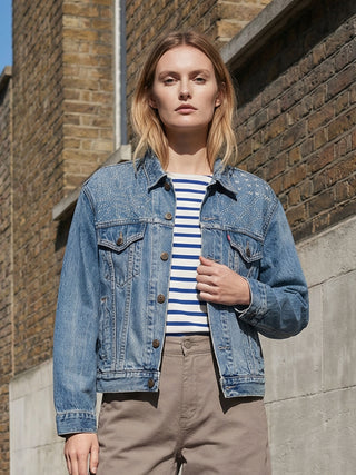 Woman wearing a denim jacket and striped shirt standing against a brick wall.