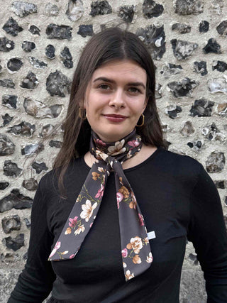 A woman with dark hair wearing a black long-sleeve top and a dark floral neck scarf, standing in front of a textured stone wall.