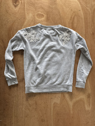 A light gray heather crewneck sweatshirt with intricate white floral lace appliques on the upper back, laid flat on a wooden surface.