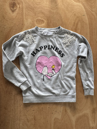 A light gray crewneck sweatshirt featuring a pink heart graphic with Snoopy and a yellow star, the word 'HAPPINESS' printed above, and white lace appliques on the shoulders, laid flat on a wooden surface.