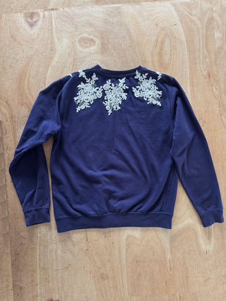A navy blue long-sleeve sweatshirt with intricate silver floral embroidery across the upper back, laid flat on a light wood background.