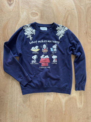 A navy blue crewneck sweatshirt featuring a Peanuts graphic print with Snoopy and Woodstock illustrations and the text 'What makes me happy' on the front, finished with white floral applique on the shoulders, laid flat on a light wood background.