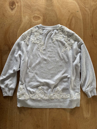 A light gray sweatshirt with intricate white floral lace embroidery on the shoulders and a matching lace band at the hem, laid flat on a wooden surface.