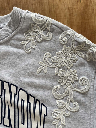 A close-up of a light gray sweatshirt featuring intricate white floral lace applique on the shoulder and collar area, with part of a navy and white collegiate-style letter graphic visible.
