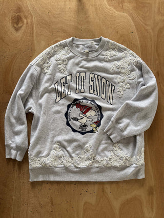 A light gray crewneck sweatshirt featuring 'LET IT SNOW' text and a Snoopy graphic on the front, embellished with white lace appliques along the shoulders and hem, laid flat on a wooden surface.