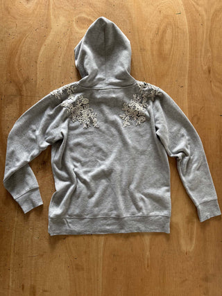 A light gray hooded sweatshirt laid flat on a wooden surface, featuring textured floral lace applique patches on the shoulders.