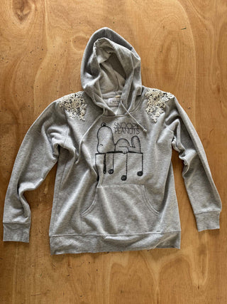 A light gray pullover hoodie with a black line art graphic of two figures and the word 'SNOOPY' across the chest, laid flat on a wooden surface. The sleeves feature decorative sequined embellishments.