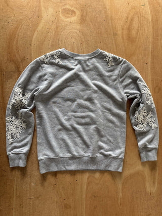 A light gray crewneck sweatshirt with intricate white lace applique detailing on the shoulders and both sleeves, laid flat on a wooden surface.