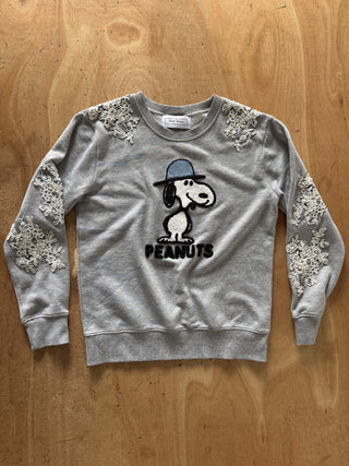 A heather gray crewneck sweatshirt featuring a Snoopy graphic on the front with 'PEANUTS' text and white lace applique on the shoulders and sleeves, laid flat on a wooden background.