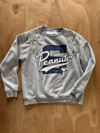 A light gray crewneck sweatshirt featuring a navy blue and white 'Peanuts' graphic on the front, embellished with white lace appliques on the shoulders, laid flat on a wooden surface.