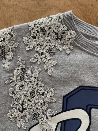 Close-up of a heather gray crewneck sweatshirt featuring intricate white floral lace applique across the upper chest and shoulder area, with a navy and white graphic letter partially visible.
