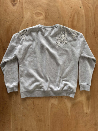 A light gray heather crewneck sweatshirt with intricate white floral lace applique on the shoulders, laid flat on a wooden surface.