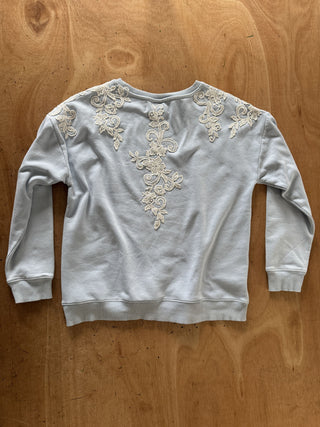A light gray sweatshirt with intricate cream-colored floral embroidery on the back, laid flat on a wooden surface.