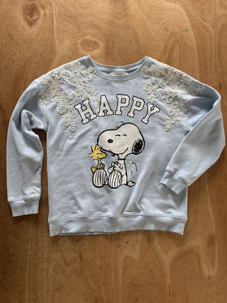 A light blue crewneck sweatshirt featuring a Snoopy and Woodstock graphic on the front with 'HAPPY' written above them, laid flat on a wooden surface.