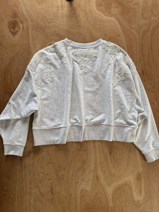 A light heather gray cropped sweatshirt with intricate white floral lace applique on the shoulders and upper back, laid flat on a wooden surface.