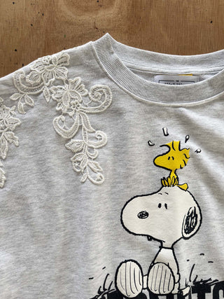 A light gray heather crewneck sweatshirt featuring a graphic of Snoopy and Woodstock, with white lace applique detailing on the upper left shoulder, laid flat on a wooden surface.