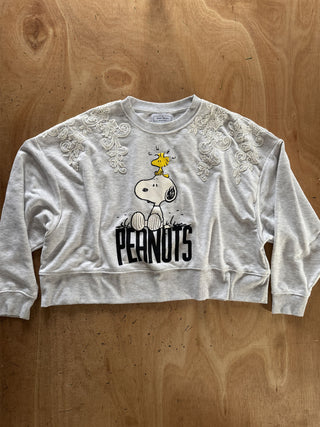 A light gray heather crewneck sweatshirt featuring a Snoopy graphic with the word 'PEANUTS' on the front, laid flat on a wooden surface.
