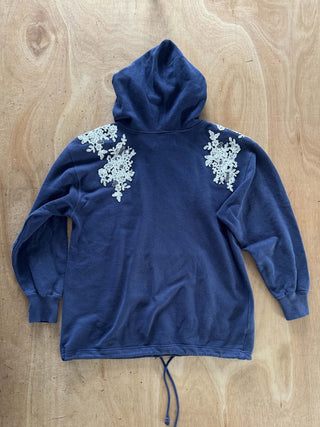 A navy blue hooded sweatshirt with intricate white floral lace appliques on the shoulders, laid flat on a light brown wooden surface.
