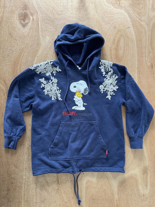 A navy blue hooded sweatshirt featuring Snoopy and Woodstock on the front, with white floral lace appliques on the shoulders and a red 'SNOOPY' text print. The hoodie is laid flat on a light wood grain surface.