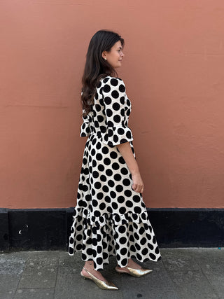 A woman wearing a black and white polka dot midi dress with ruffled sleeves and a tiered hem, standing against a terracotta-colored wall.