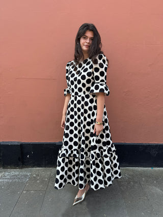 A woman wearing a black and white polka dot midi dress with puff sleeves, standing against a terracotta-colored wall.