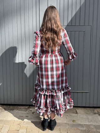 A woman viewed from behind wearing a red, white, and black plaid midi dress with ruffled sleeves and a tiered ruffle hem, standing in front of a gray wooden door.
