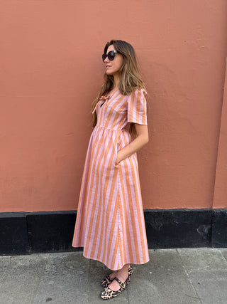 A woman wearing a peach and white vertically striped midi dress with short sleeves, standing against a textured terracotta wall.