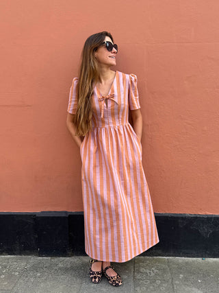 A woman wearing a pink and white vertically striped midi dress with puff sleeves and a bow at the neckline, standing against a textured terracotta wall.