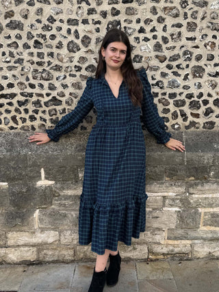 A woman wearing a dark blue and black plaid midi dress with long sleeves and a ruffled hem, standing outdoors against a rustic stone wall.