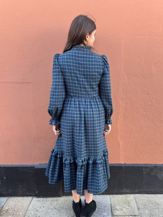 A woman viewed from behind wearing a dark blue and black plaid midi dress with puff sleeves and a ruffled hem, standing against a textured terracotta wall.